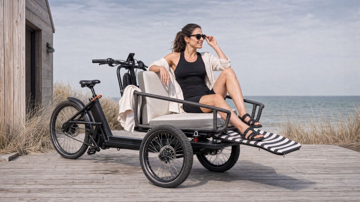 The Trike parked on a seaside boardwalk with its flat-bed module set up as a lounge seat, a rider relaxing on the cushions in the sun.