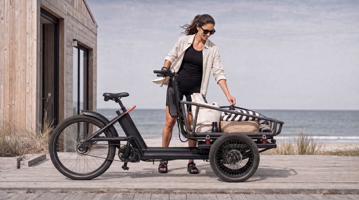 A rider lifting a canvas bag into the Trike's cargo area on a boardwalk beside a wooden cabin, with the sea in the background.